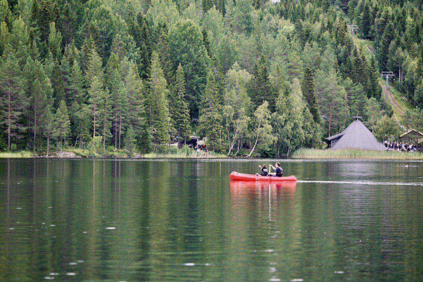 A red canoe with two paddlers on a still lake ringed by conifer forest