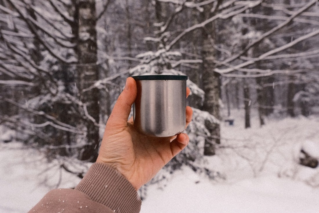 A hand holding a metal camp mug in a snowy birch forest